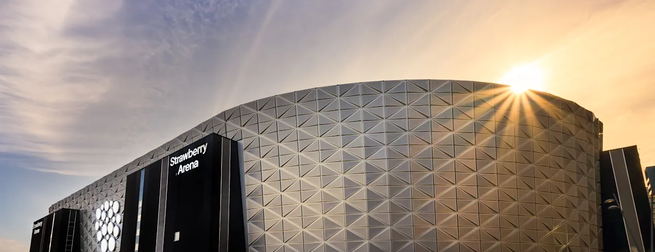 The Strawberry Arena building at sunset, with the sun shining brightly behind it. The building has a modern, geometric facade.