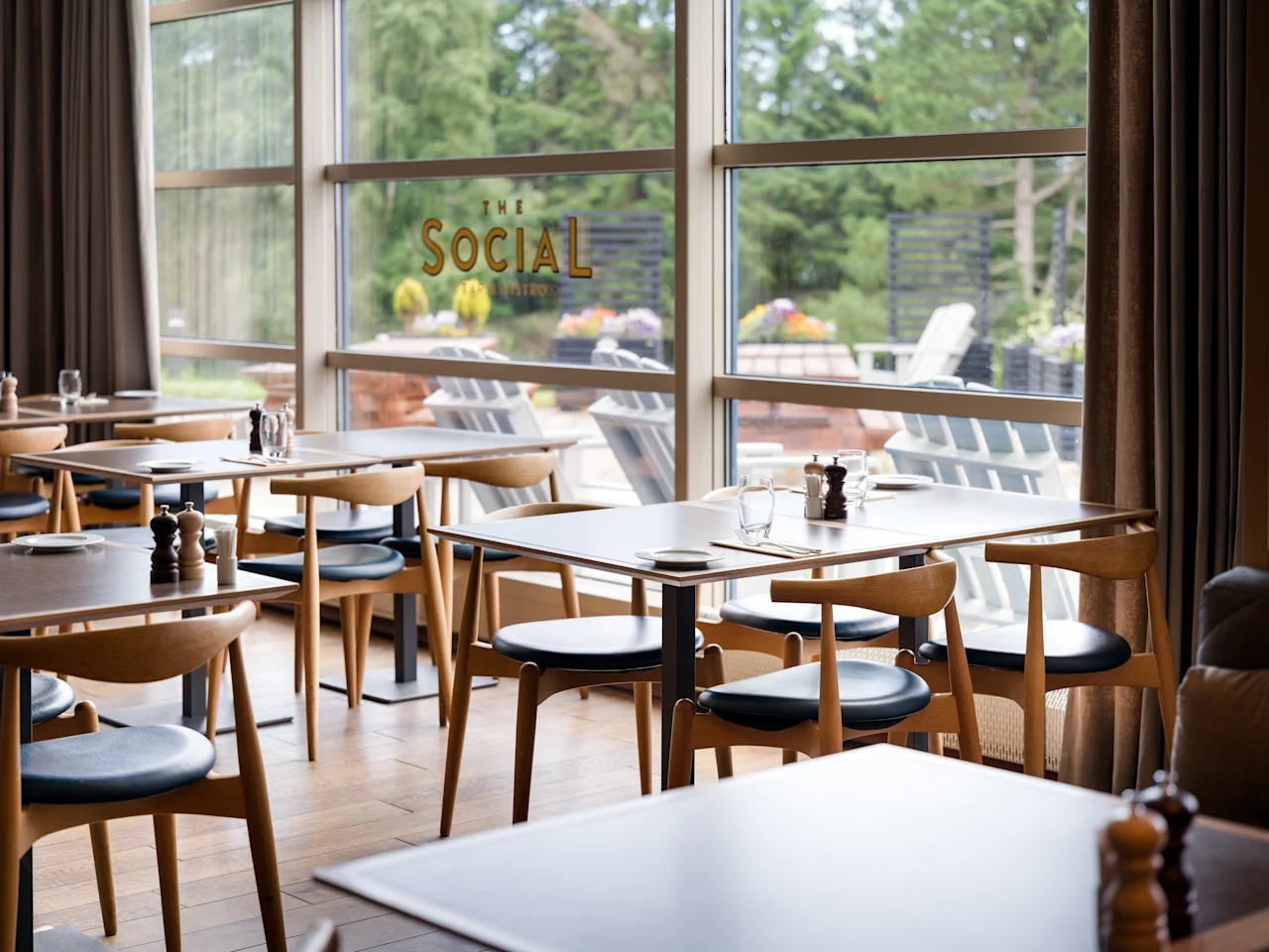 Moderni ravintola, jossa on katetut puupöydät ja tuolit suurten ikkunoiden äärellä ja näkymä vihreyteen.
