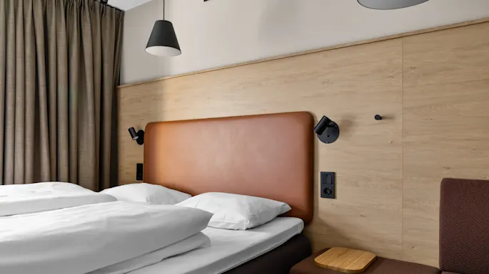 A bed with white linens and a brown headboard is flanked by black reading lamps. It's adjacent to brown curtains, a wooden wall panel, and a cushioned bench.