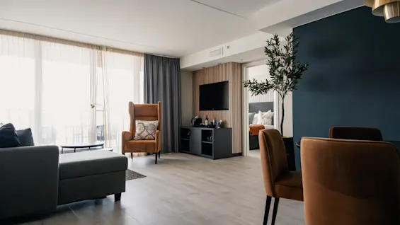 En stue har en brun lenestol ved siden av en sofa, vendt mot en TV på veggen. Et hjørne er dekorert med en plante, og en seng er synlig gjennom døren.