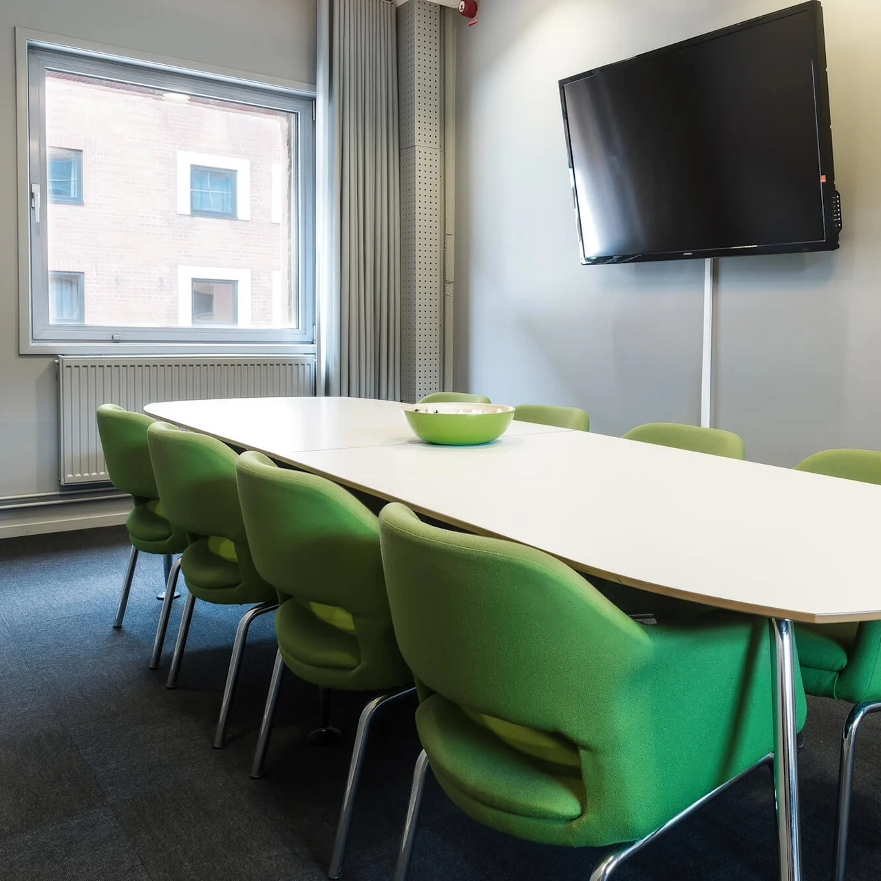 A long white table surrounded by green chairs is positioned under a wall-mounted TV in a meeting room with a window overlooking a brick building.
