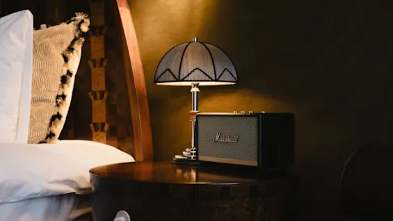 En Marshall-høyttaler hviler på et tre-nattbord ved siden av en lampe i et svakt opplyst rom.