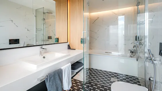 A sleek bathroom features a large mirror above a white sink with a towel below, adjacent to a glass-enclosed bathtub and shower; marbled walls and black tiles provide a modern aesthetic.