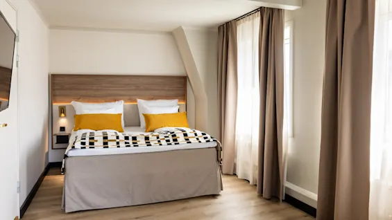 Viihtyisä hotellihuone mukavalla sängyllä, keltaisilla tyynyillä ja lämpimällä valaistuksella, valmiina rentouttavaan oleskeluun.