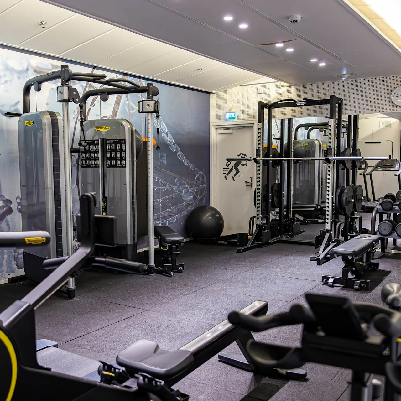 A gym features exercise machines and weights; some machines face a mirror on the wall. A clock and an outlined figure are visible above a door. 