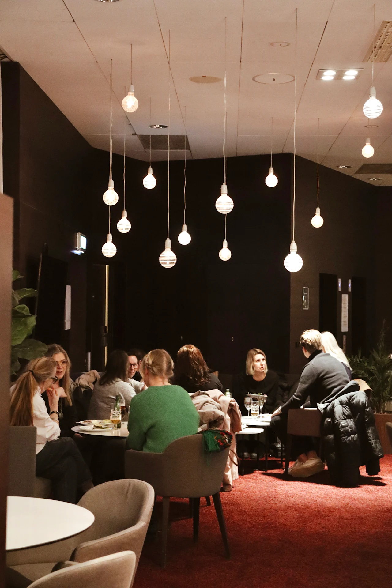 People sit at round tables, engaged in conversation and dining in a warmly-lit restaurant with hanging globe lights and red carpeting.