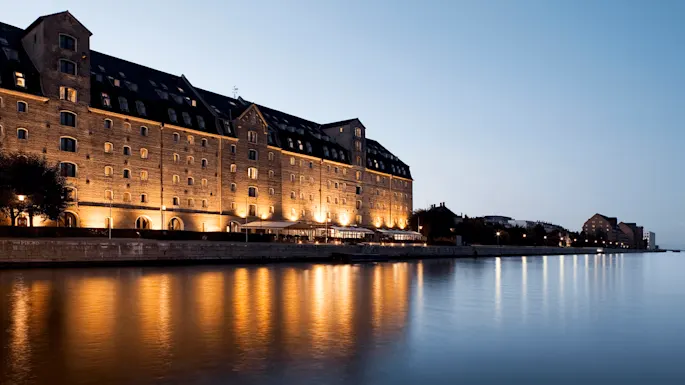 A large, illuminated brick building stands beside a calm waterfront at dusk, reflecting warm lights on the water. The sky is clear and transitioning to night.