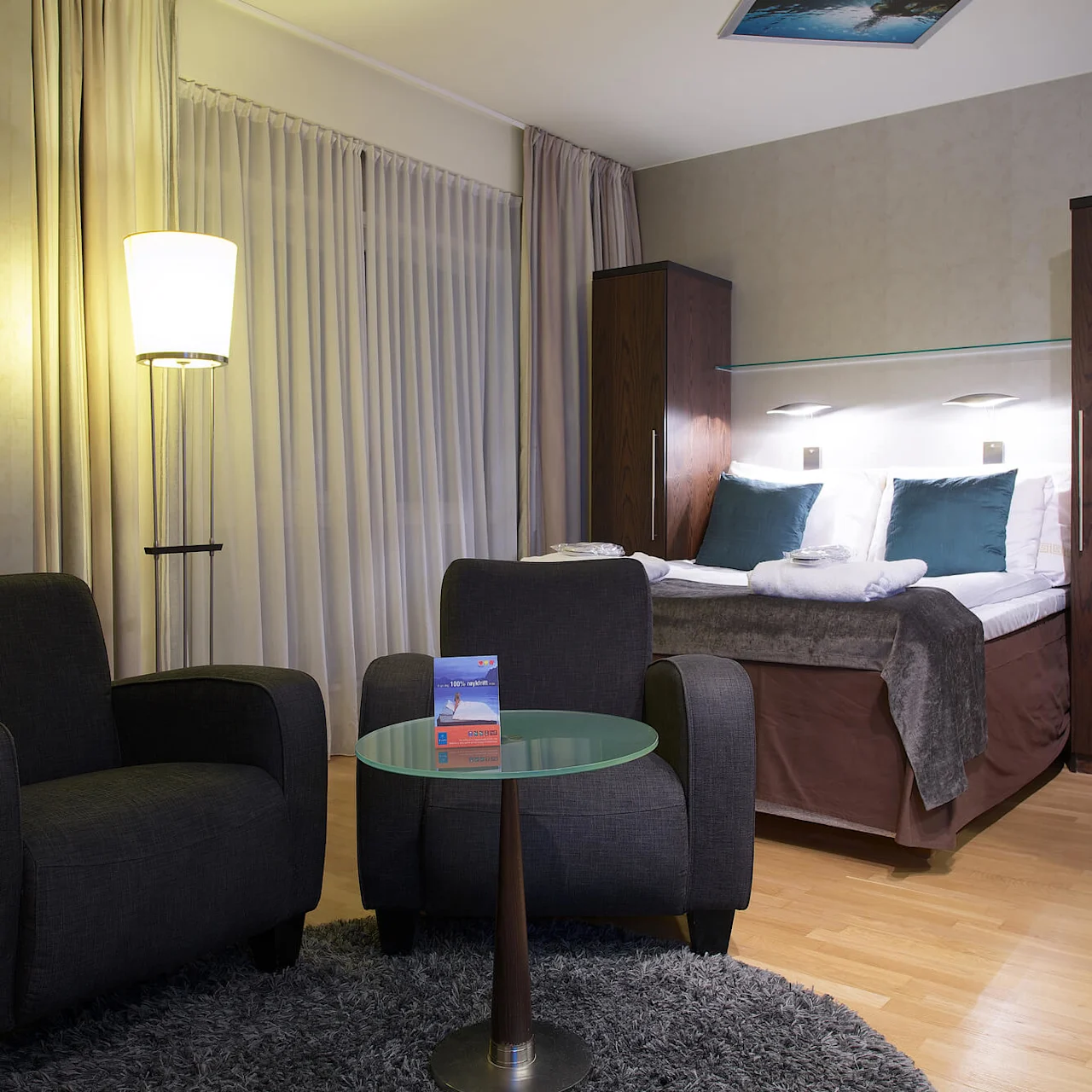 A well-lit modern hotel room with two armchairs, a glass table, and a bed in the background, setting a cozy ambiance.