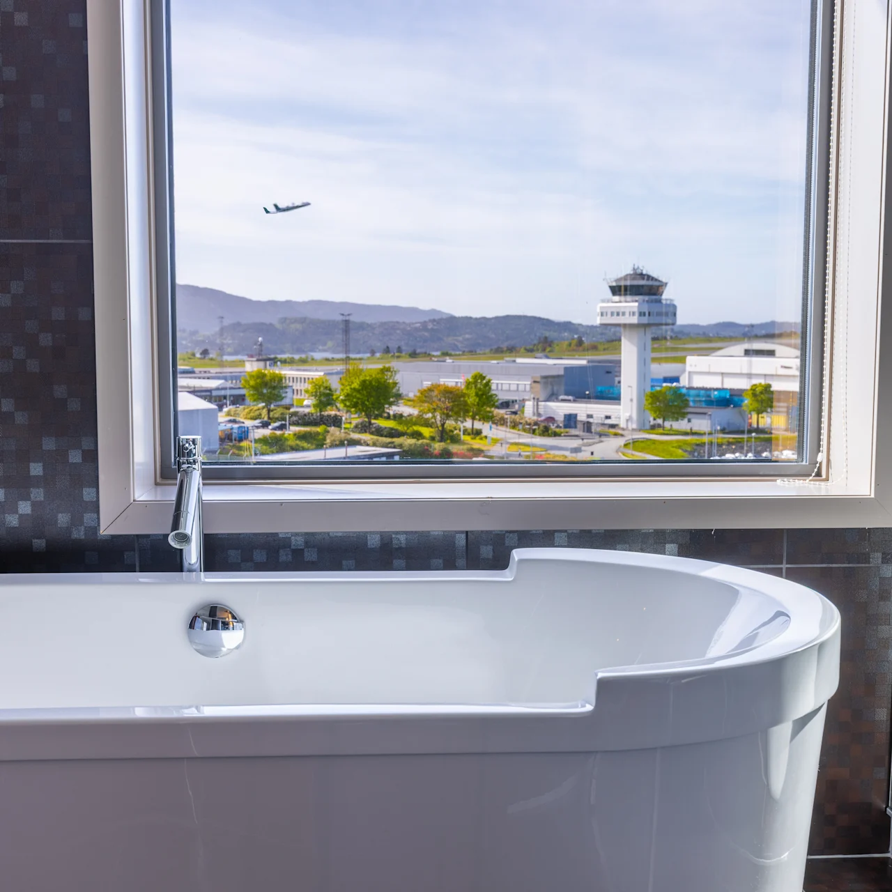 A white bathtub sits inactive in a modern bathroom. Through the window, an aircraft flies above an airport with a control tower surrounded by distant hills.