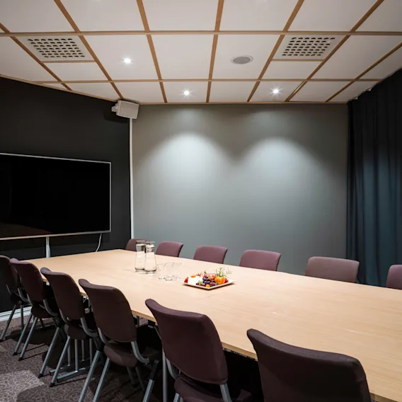 Et stort konferansebord i tre omgitt av stoler, med glasskanner og et fruktfat. Montert skjerm på en mørk vegg. Myk belysning, teppebelagt gulv, moderne atmosfære.