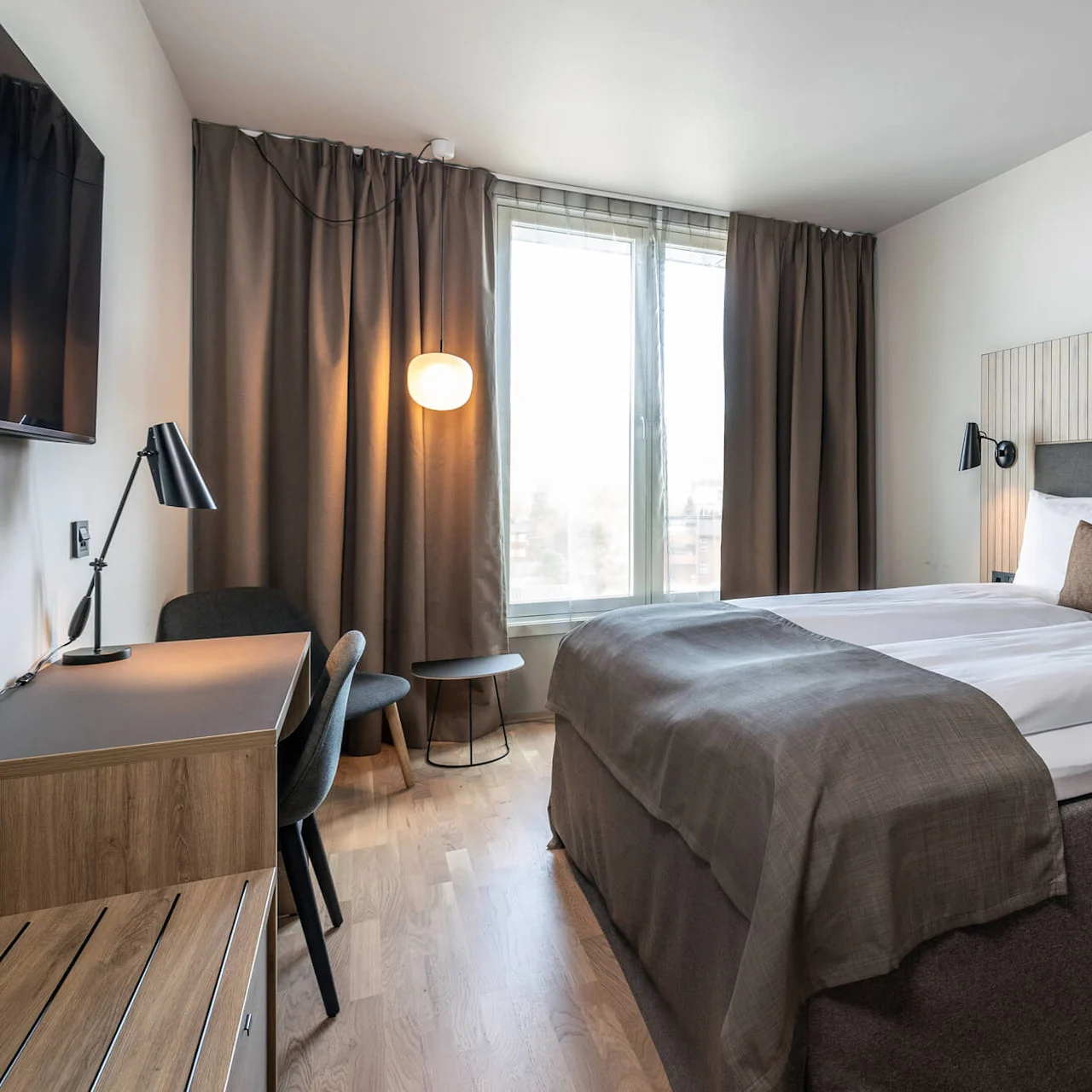 Et pænt indrettet hotelværelse har en stor seng med hvide lagener og brune detaljer, flankeret af moderne lamper. Et skrivebord med stol står i nærheden, med bløde gardiner ved et vindue.