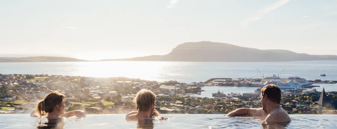 Folk slapper af i en infinitypool med udsigt over en solrig kystby, bjerge og havet.