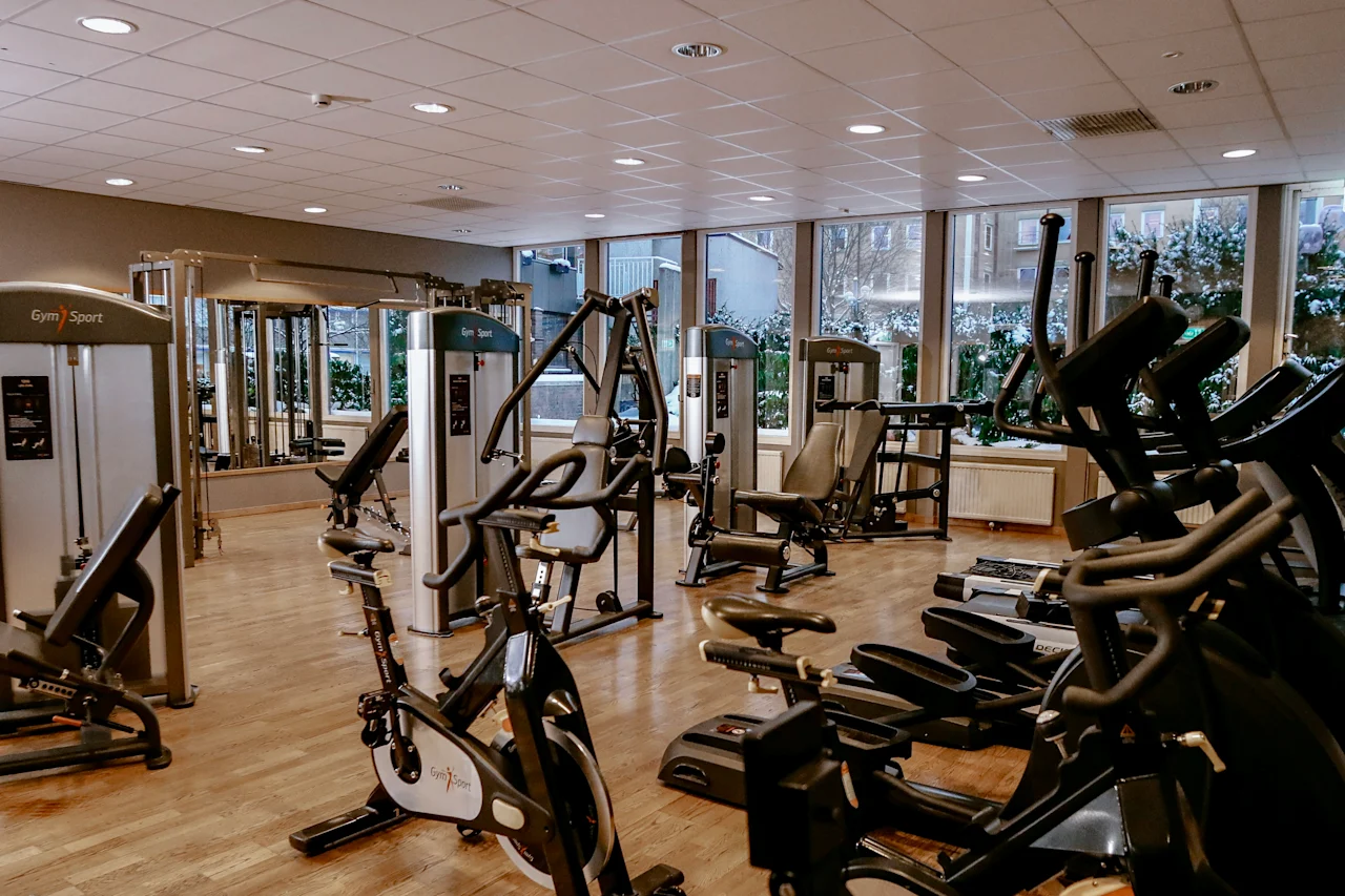 A gym with various exercise machines stands empty. The space is well-lit with large windows, reflecting a clean and organized environment. Text: "Gym|Sport".