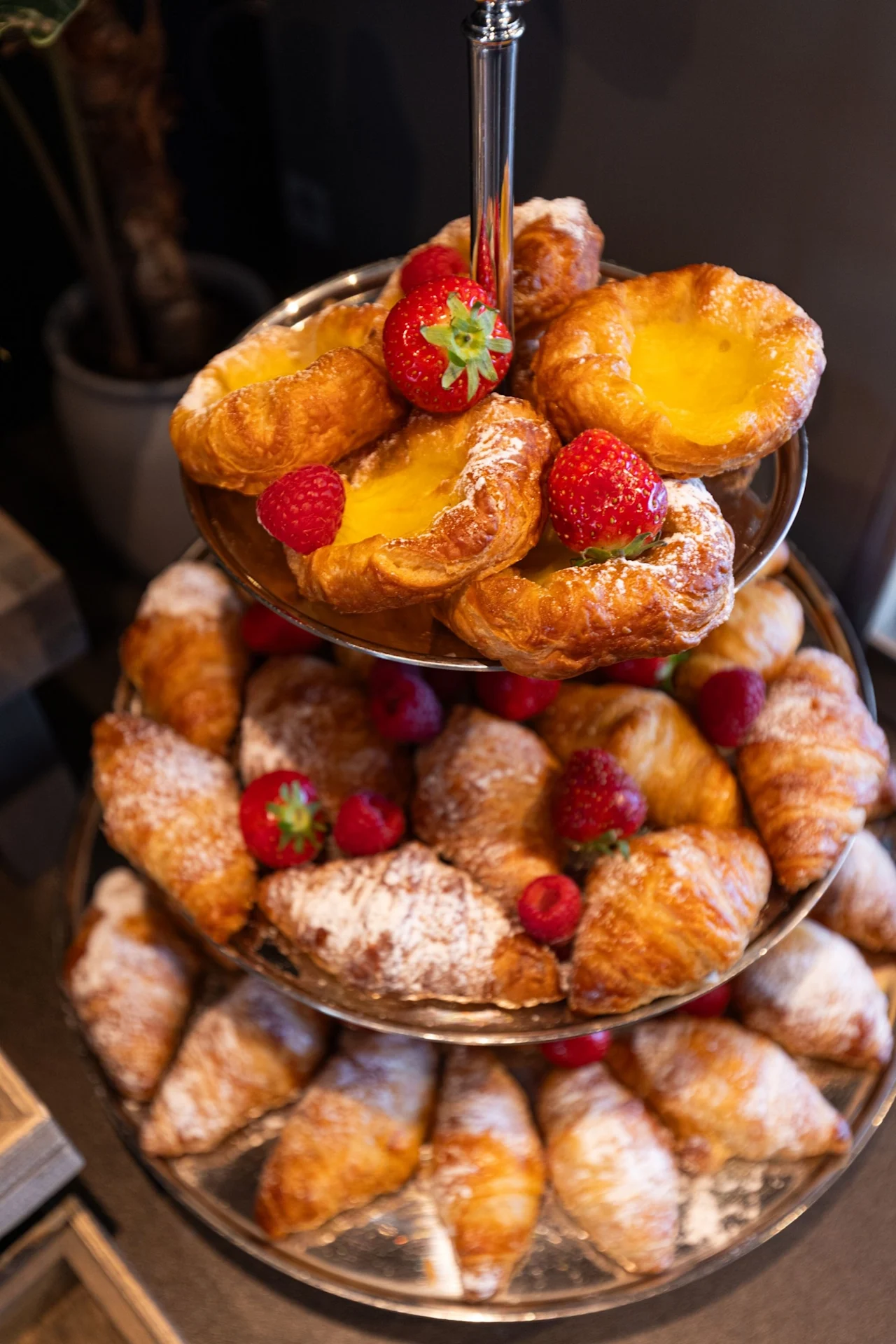 Bakverk på et etasjefat, med croissanter og sitronterter toppet med jordbær og bringebær, dekorativt arrangert på et bord.