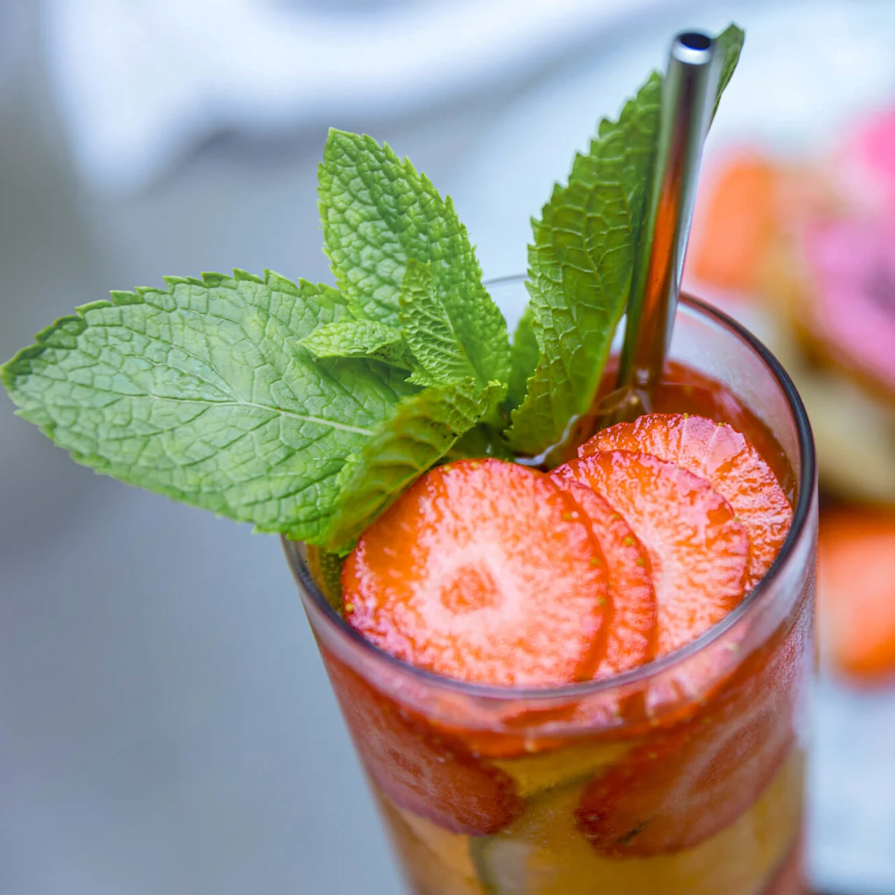 A glass of iced beverage with sliced strawberries and mint leaves is topped with a metal straw. It sits on a table with a blurred dessert in the background.