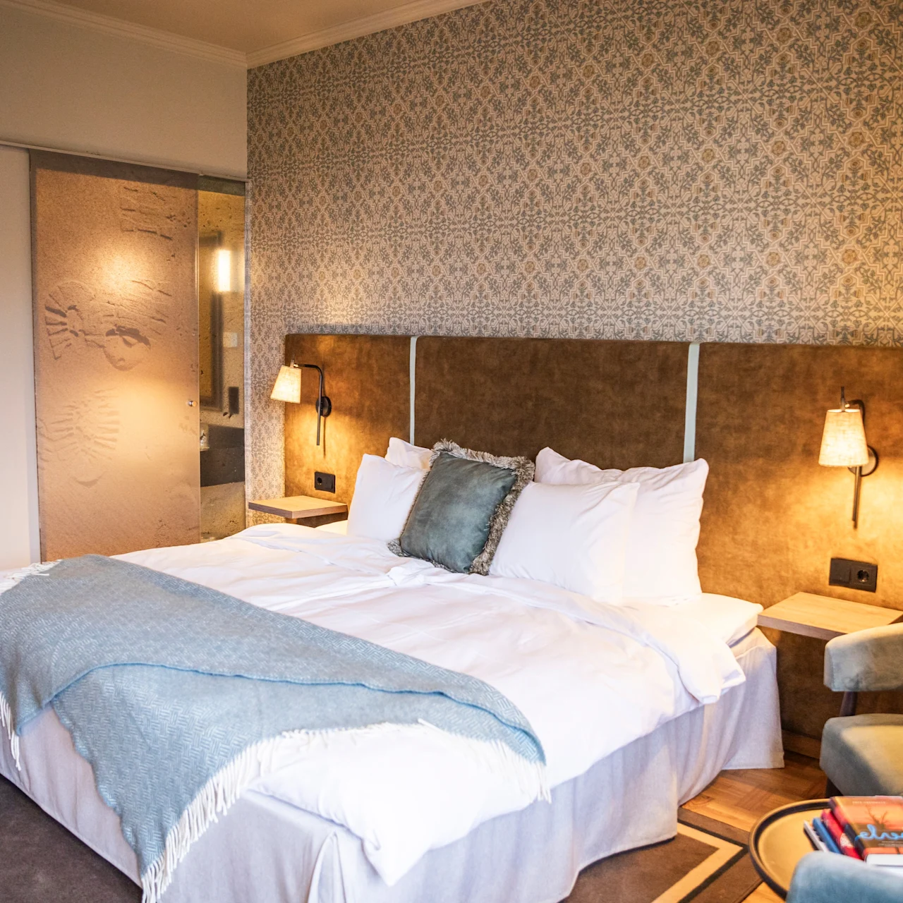 A large bed is covered with white linens and a blue blanket, situated against a patterned wallpaper in a warmly lit bedroom with side tables and a cushioned chair.