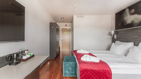 A comfortable hotel room featuring a bed with a red throw and bathrobes, a desk with a TV, and an exit sign on the door.