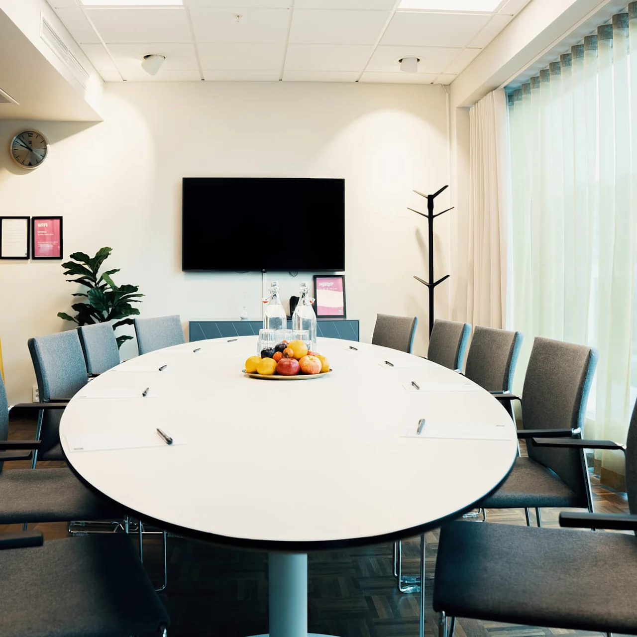 Konferensbord har papper, pennor och fruktskål; omges av stolar. Bakgrund: dekorativa inomhusväxter, klocka, gardin, inramade meddelanden. Vägg har TV. Stora fönster släpper in ljus.