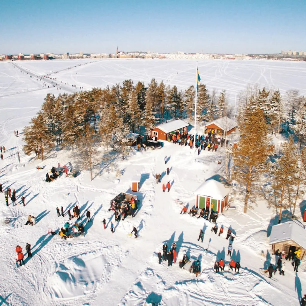 Luftfoto af vinteraktiviteter på sneklædt ø, Gråsjälören i Luleå, med frossen sø og by i baggrunden.