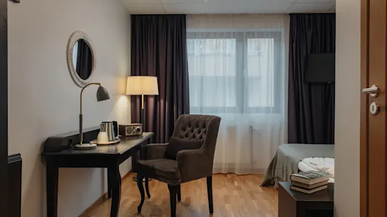 Viihtyisä hotellihuone, jossa on työpöytä, vedenkeitin, mukava nojatuoli ja sänky, jonka yöpöydällä on kirjoja.