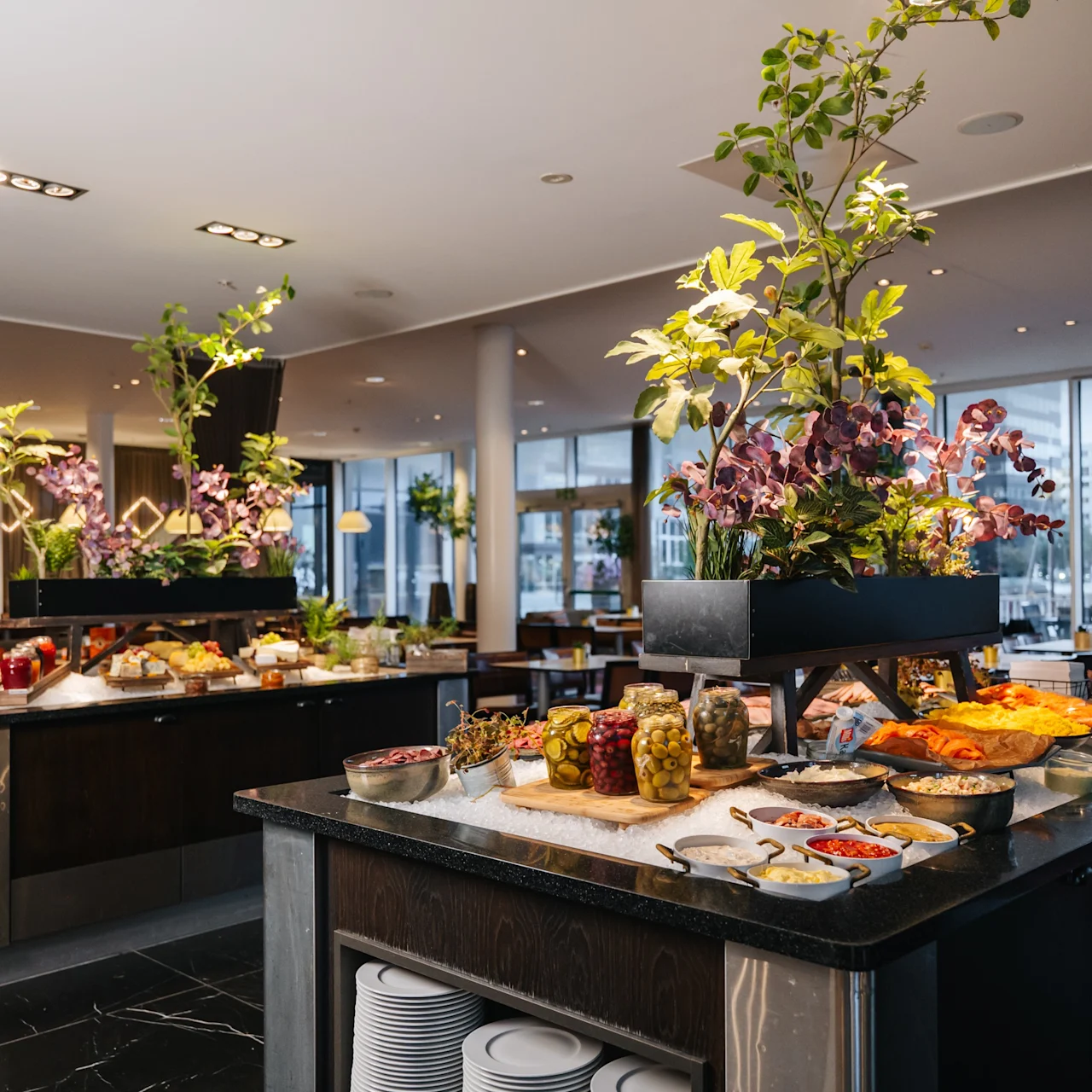 Buffet tables are adorned with various dishes and decorative flowers, set in a spacious dining area with large windows and modern lighting.