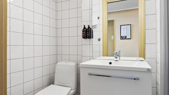 Lyst hotellbaderom med hvite fliser, toalett, servant med skuffer og et speil.