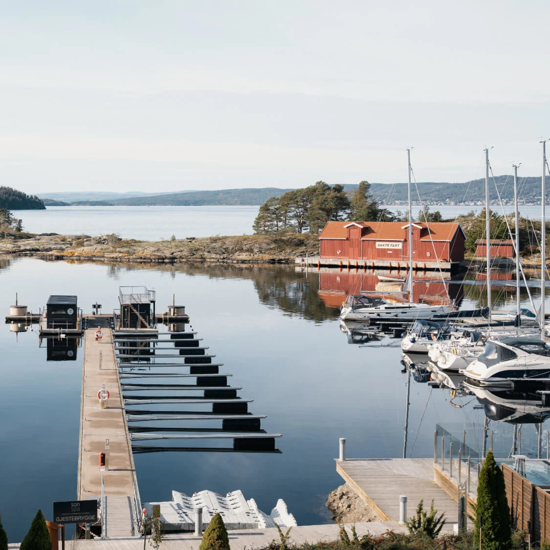 Båter fortøyd ved en marina, omgitt av rolig vann og steinete landskap. Et rødt båthus med skilt: "Sørenga", "Kafé & Bar", "Gjesteplass", og "GjesteBrygge."
