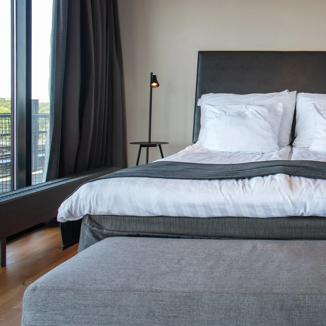 A neatly made double bed with white pillows and bedding is flanked by matching bedside lamps. It faces a tall window with dark curtains in a modern bedroom.