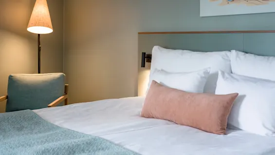A neatly made bed with white and beige pillows stands in a serene room, accompanied by a floor lamp and modern armchair. A colorful painting decorates the muted walls.