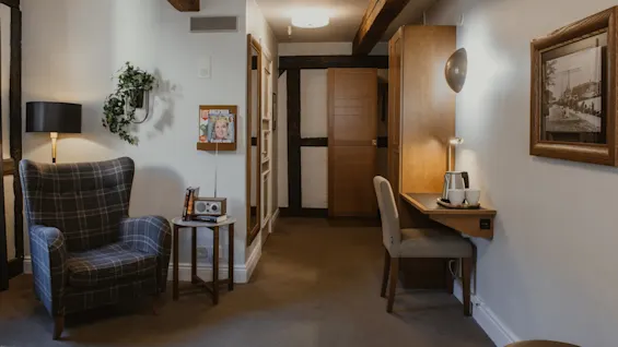 Et hyggeligt hotelværelse med en ternet lænestol, et læsehjørne med bøger og et skrivebord med en elkedel. Magasiner på væggen, UT Evigt ung.