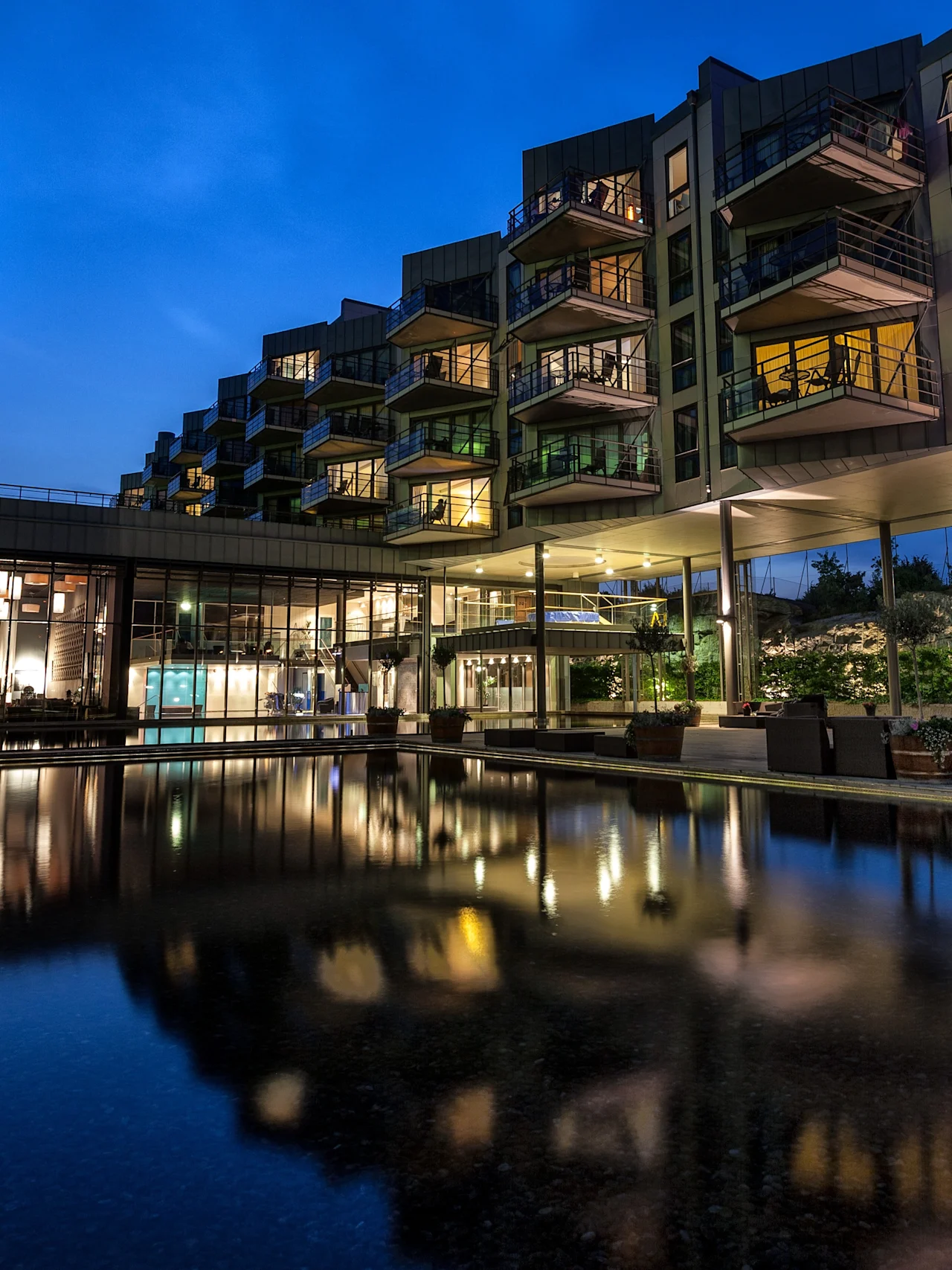A modern building with illuminated balconies reflects in a calm water feature at dusk, surrounded by green trees and outdoor dining areas under a deep blue sky.