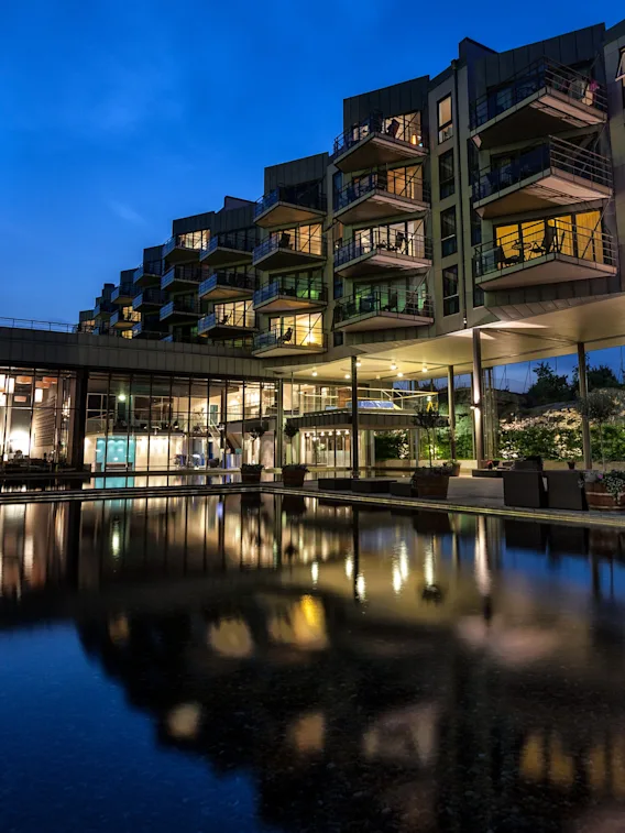 En modern byggnad med upplysta balkonger reflekteras i en lugn damm i skymningen, omgiven av gröna träd och utomhusrestauranger under en djupblå himmel.