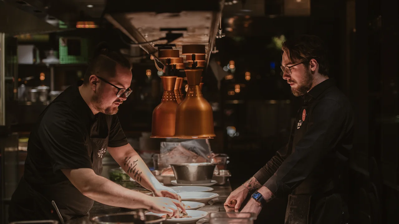 Two chefs meticulously preparing dishes in a professional kitchen under warm lighting.