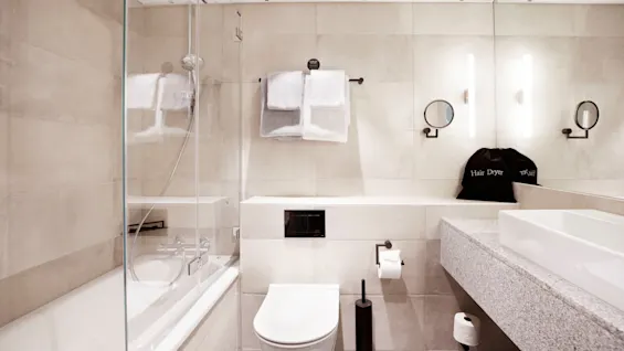 Modern bathroom featuring a white toilet, bathtub with glass screen, and a countertop sink. Towels hang above the toilet. A hair dryer bag is visible. Soft lighting enhances the clean, tiled space.