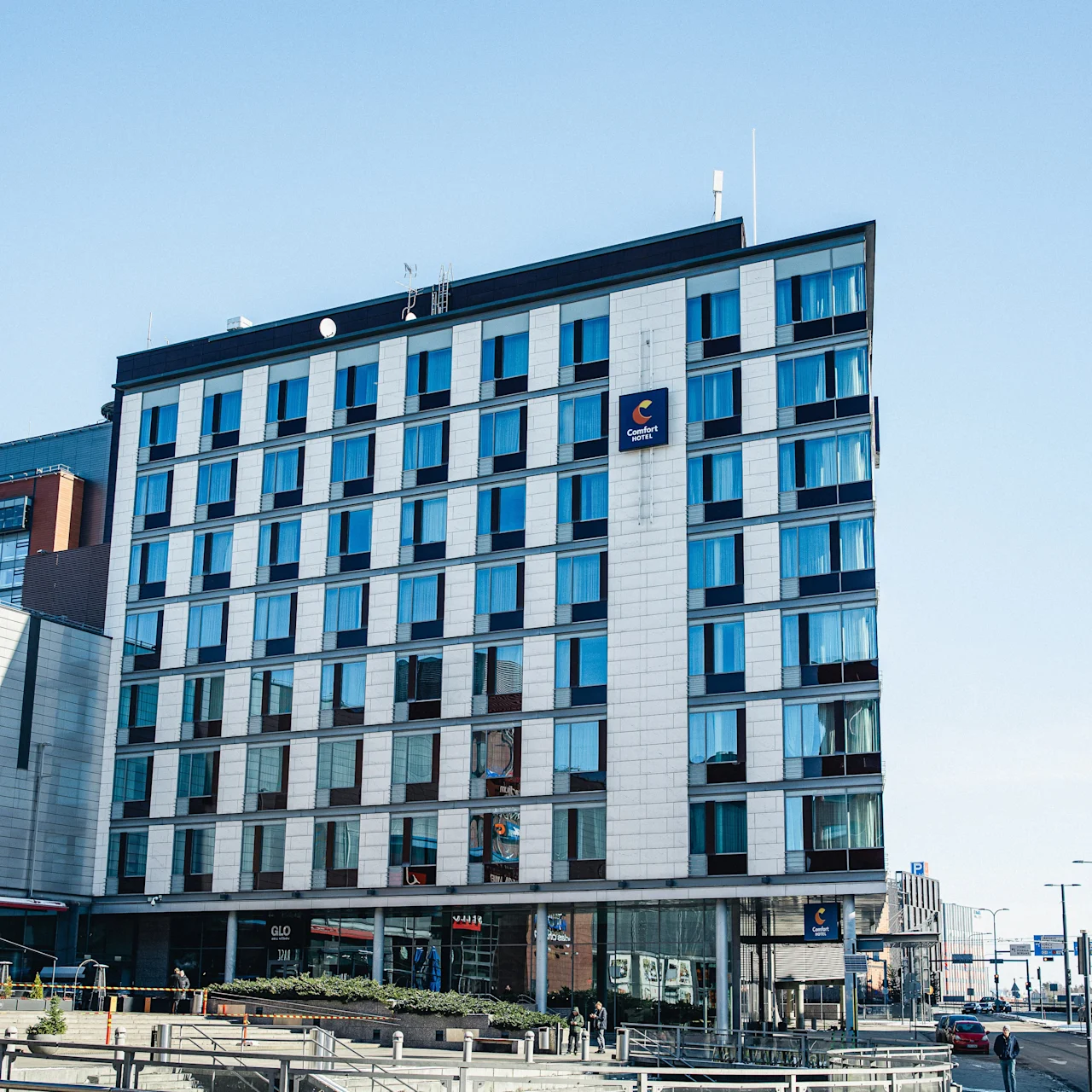 Hotel building stands tall, displaying "Comfort Hotel" signage against a clear blue sky, surrounded by urban landscape with plant boxes and pedestrians in the foreground.