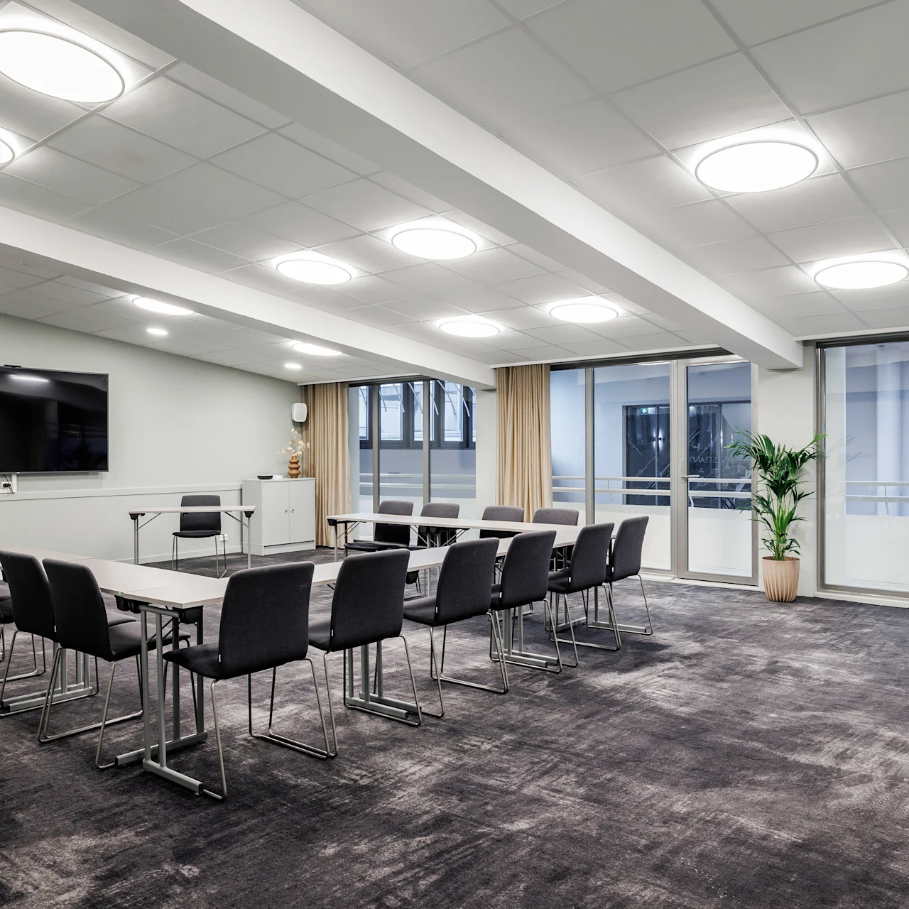 Conference room with rectangular tables and black chairs arranged in a U-shape, facing a large wall-mounted screen. Windows and potted plants provide a modern, well-lit environment.