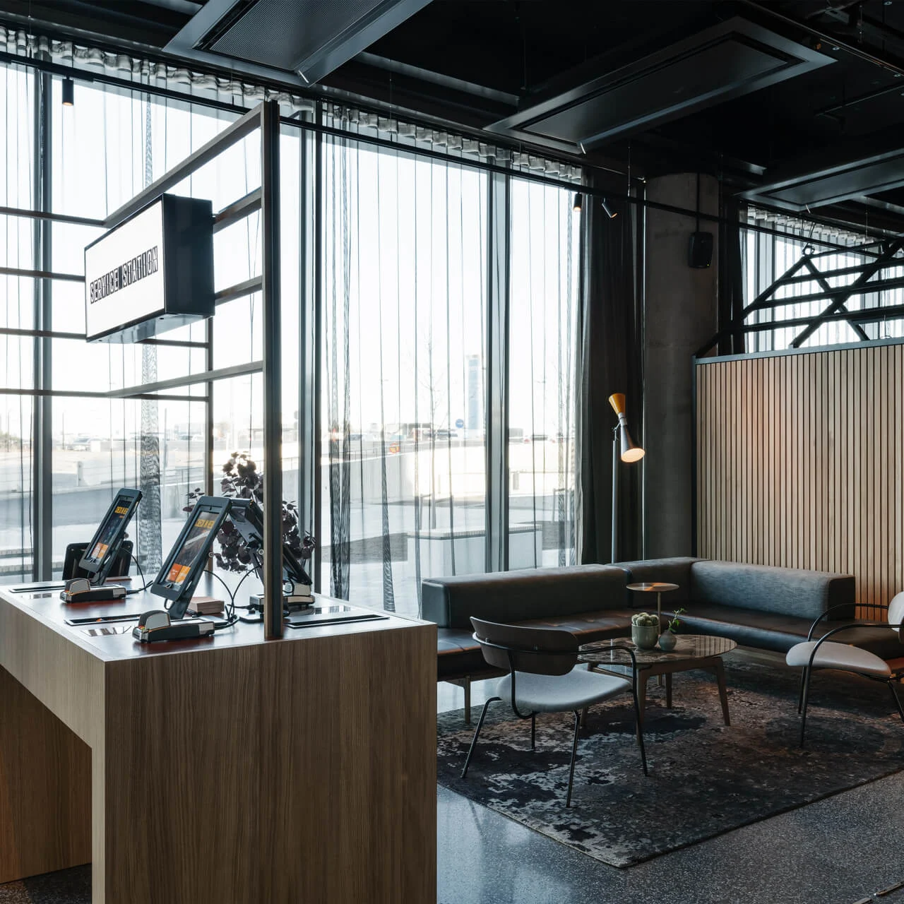 Modern reception area with two self-service kiosks on a wooden desk, in a spacious room with large windows and stylish seating.