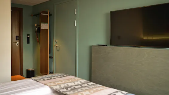 En seng med et mønstret tæppe er i et pænt hotelværelse, vendt mod et vægmonteret TV. En dør, knagerække og mørk dørkarm indrammer rummet.