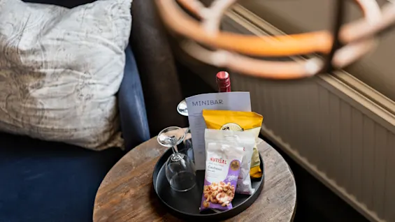 Et minibararrangement på et træbord ved siden af en lænestol, med en flaske, et vinglas og snackposer.