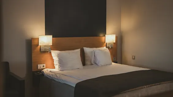 Viihtyisä hotellihuone siististi pedatulla sängyllä, valaistuna lämpimillä yöpöytälampuilla, kutsuen rentoutumaan.