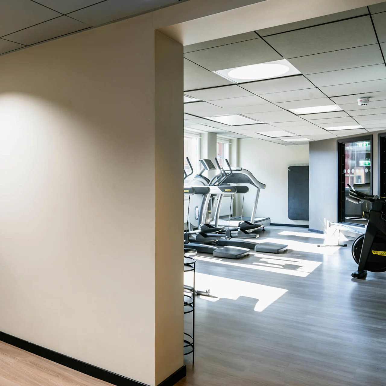 Håndvaskområde med vandhane og bordplade, ved siden af et fitnesscenter fyldt med løbebånd, motionsmåtter og stationære cykler, oplyst af naturligt lys gennem store vinduer.