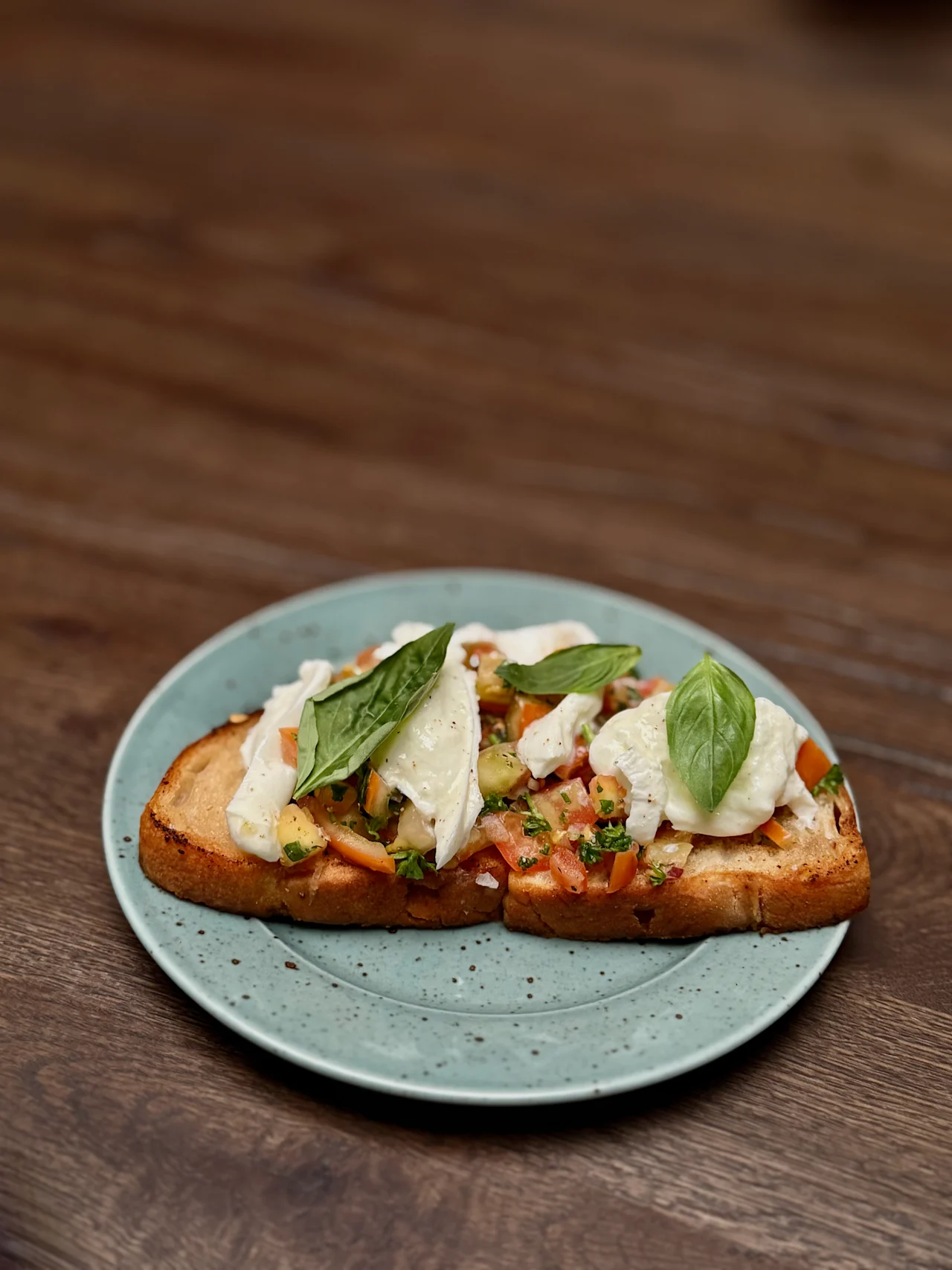 A slice of toasted bread topped with diced tomatoes, mozzarella slices, and basil leaves rests on a speckled green plate, placed on a wooden table.