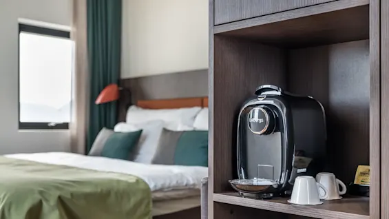 A coffee machine labeled "Löfbergs" rests on a wooden shelf next to two cups, tea bags, and a tidy bed with green bedding in a well-lit room.