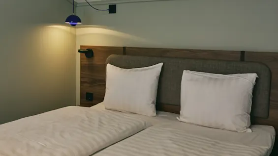 A hotel room featuring a bed with white pillows and striped bedding, illuminated by a wall-mounted lamp.