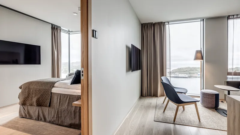 En moderne hotelsuite med soveværelse og opholdsstue, med store vinduer med udsigt over en naturskøn havnefront.