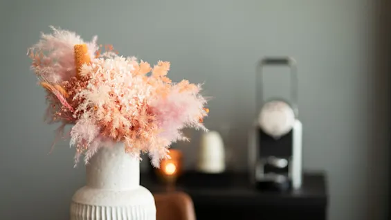 En hvit vase fylt med et vakkert arrangement av tørkede rosa og oransje blomster, mot en myk grå bakgrunn.