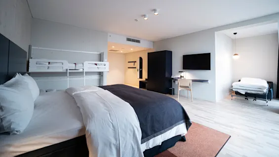 A large bed with white and dark bedding is in a spacious modern hotel room, featuring a mounted TV, desk, chair, and a pull-out bed in a nook.