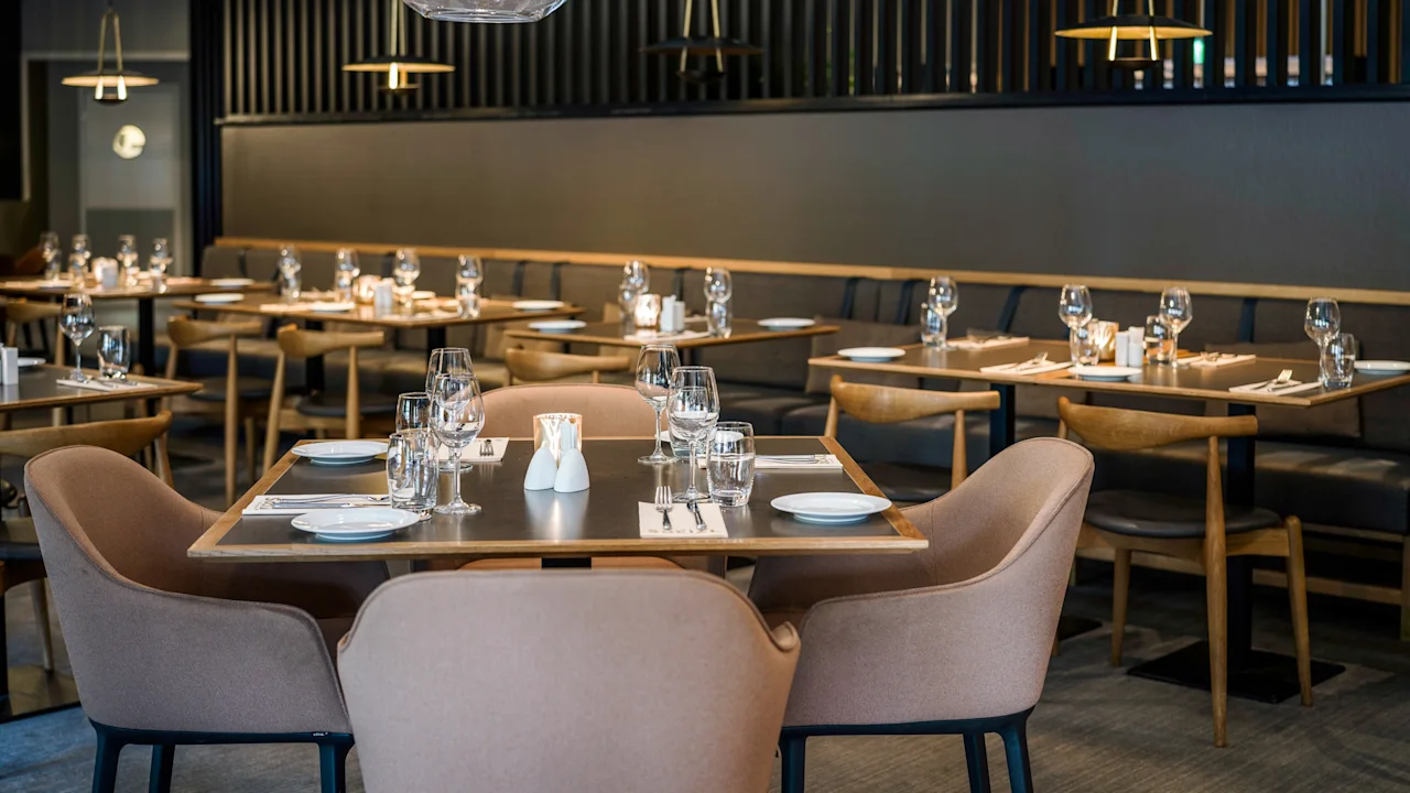 En hyggelig, moderne restaurant med pænt dækkede borde, blød belysning og elegante stole. Vinglas og bestik er arrangeret, hvilket skaber en indbydende atmosfære.