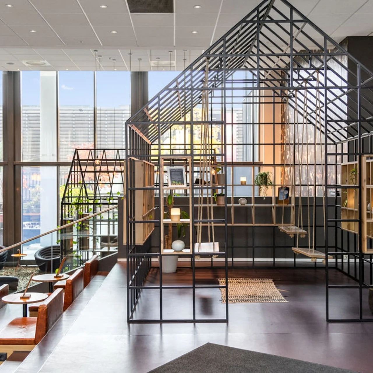 A metal-framed structure resembling a house stands indoors with shelves and seating, surrounded by modern furniture and large windows allowing natural light. No text is visible.