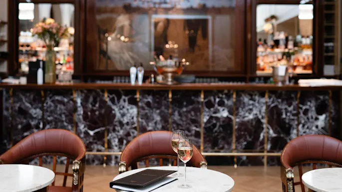 Et glass vin står på et marmorbord, flankert av elegante stoler og en stilig bar i bakgrunnen.